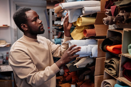 Young African American male leatherworker choosing suitable color of suede or leather textile for...