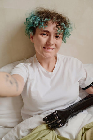 Smiling curly young woman with prosthetic arm taking selfie in bed