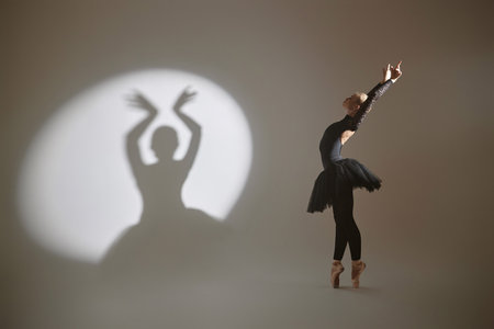 Ballerina posing on tiptoes with her arms raised and back bent back, her shadow cast on wall on leftの写真素材
