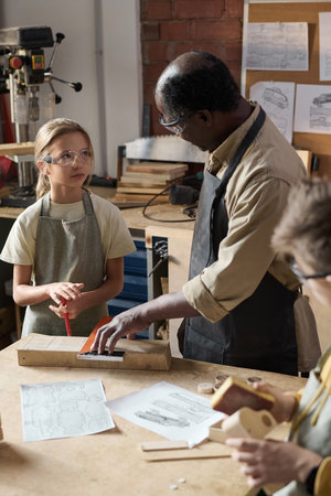 Black senior man teaching carpentry class for children in workshop and looking at little girl
