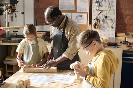 Portrait of senior craftsman teaching children carpentry in workshop lit by sunlight, copy spaceの写真素材