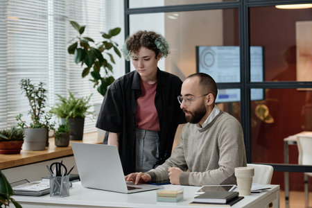 Man sitting at office desk looking at laptop screen, his young female colleague standing next to him
