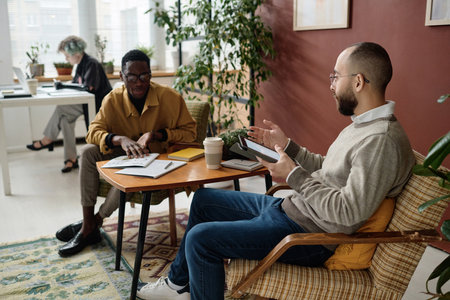 Biracial male coworkers discussing documents with infographics sitting at table in office