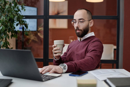 Man in casual outfit sitting at his desk in office browsing laptop with reusable coffee cup in hand
