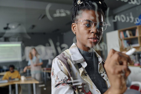 Female coder thoughtfully writing on glass wall in office, her colleagues working on backgroundの写真素材