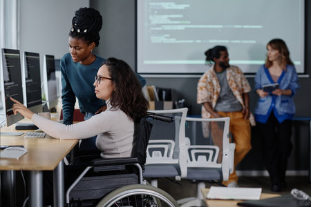 Black woman helping her colleague in wheelchair with code writing, coworkers standing on backgroundの写真素材