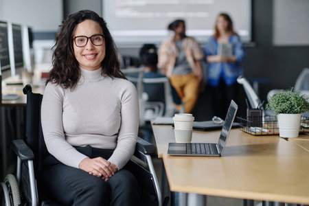 Smiling woman in wheelchair posing at workplace in office, her colleagues standing on backgroundの写真素材