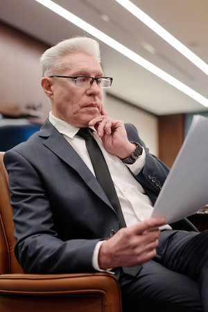Senior businessman in formalwear sitting in armchair and thoughtfully reading information on paper