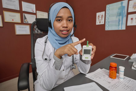 Young medical worker holding glucometer and looking at camera sitting at her workplace in clinic