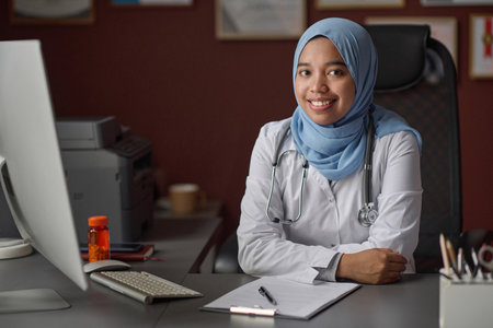 Portrait of female medical worker in hijab and uniform sitting at desk in clinic smiling at cameraの写真素材