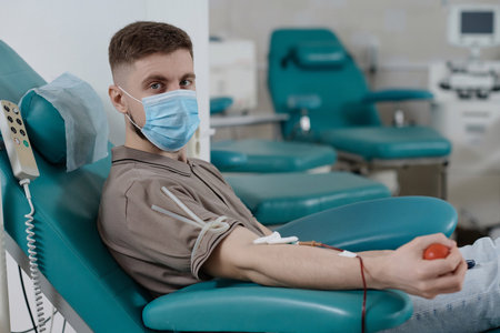 Young man in protective mask sitting in medical chair during blood donation and looking at cameraの写真素材