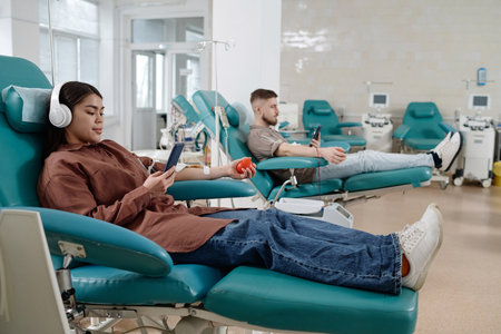Blood donors sitting side by side on medical recliner chairs in hospital and browsing smartphones
