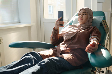 Muslim woman relaxing on medical chair and scrolling smartphone during blood transfusion in hospitalの写真素材