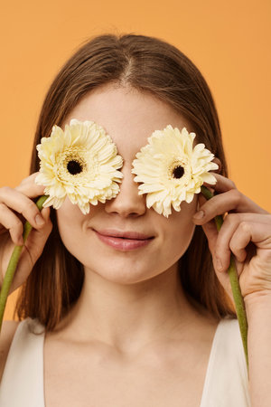 Vertical closeup studio portrait of modern young Caucasian woman covering her eyes with two gerbera flowers while posing for cameraの写真素材