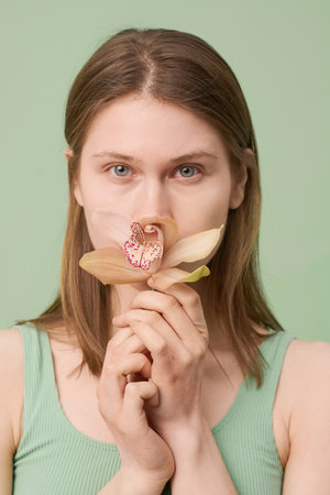 Vertical closeup studio portrait of young Caucasian woman with brown hair smelling orchid flower and...