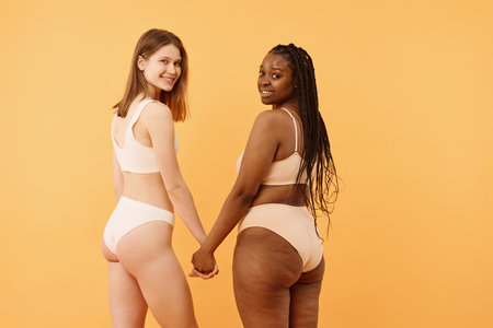 Studio shot of self-confident African American and Caucasian girls in underwear holding hands and smiling at camera, warm apricot backgroundの写真素材