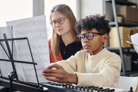 African American boy wearing eyeglasses sitting at synthesizer reading sheet music, his teacher sitting next to himの写真素材