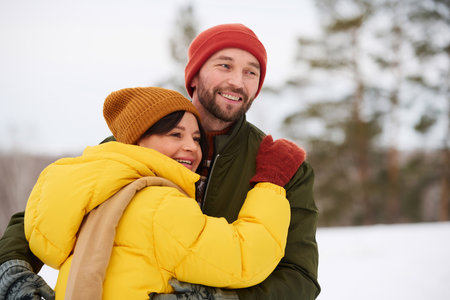 Medium shot of joyful Caucasian couple spending winter day hiking in mountains, hugging and looking awayの写真素材