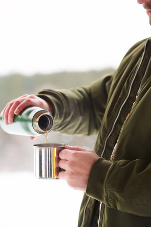 Unrecognizable tourist wearing warm clothes pouring hot tea from vacuum flask into mug on cold winter dayの写真素材