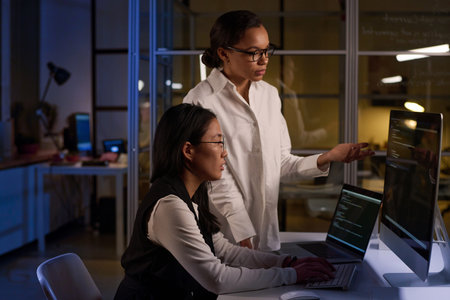 Ethnically diverse female programmers fixing code at night in modern officeの写真素材