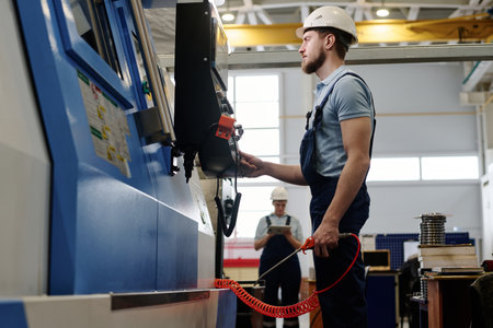 Low angle medium long side view of bearded Caucasian man wearing uniform with hardhat using CNC...