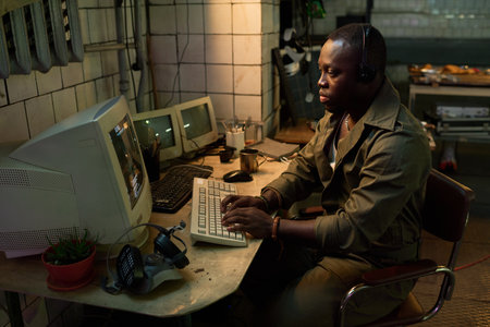 Serious African American IT specialist wearing headphones coding on old computer in abandoned...