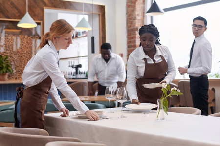 Ethnically diverse waiting staff working in modern restaurant preparing tables for guests in morning
