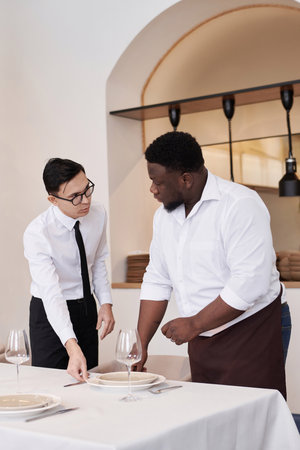 Vertical medium long shot of Asian headwaiter and African American busser discussing work while setting table in modern restaurantの写真素材