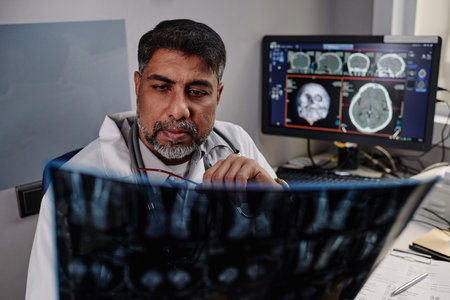 Modern mature biracial male radiographer sitting at table in his office working with brain X-ray scan imagesの写真素材