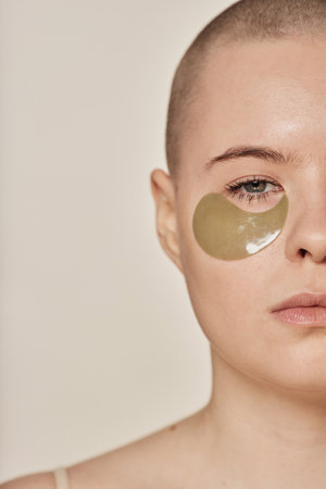 Half face studio portrait of young Caucasian woman with shaved head and with collagen eye patches looking at cameraの写真素材