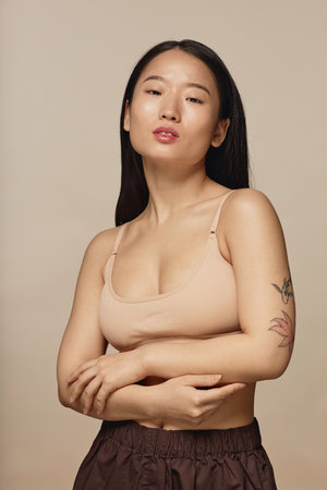 Vertical medium studio portrait of young Asian woman wearing beige bra and brown pants posing for cameraの写真素材