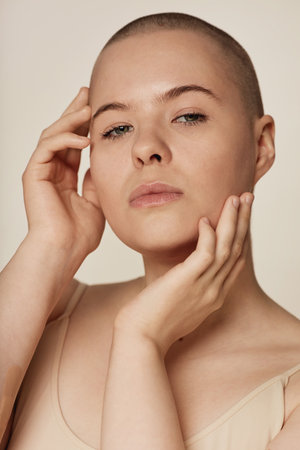 Vertical closeup studio portrait of young Caucasian woman with shaved head touching her face with hands while posing for cameraの写真素材