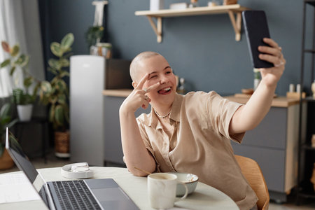 Stylish young adult Caucasian woman with shaved head sitting in kitchen posing while taking selfie on smartphoneの写真素材