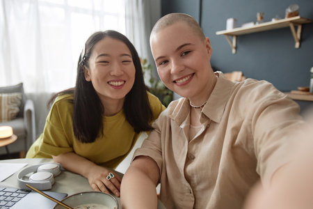 POV of joyful young Caucasian woman with shaved hand holding camera taking selfie photo with her Asian girl friendの写真素材