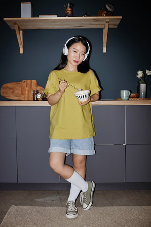 Vertical long flash shot of stylish young Asian woman in headphones wearing yellow t-shirt and denim shorts standing in kitchen eating mealの写真素材