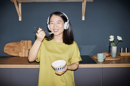 Flash portrait of joyful young Asian girl wearing yellow-green T-shirt and headphones standing against kitchen counter smiling at camera while eatingの写真素材