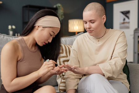 Young Asian woman sitting on couch applying nail polish on her Caucasian friends fingernails, medium...