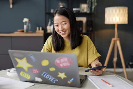 Joyful young adult Asian woman sitting at table in modern kitchen surfing Internet on laptop and smilingの写真素材