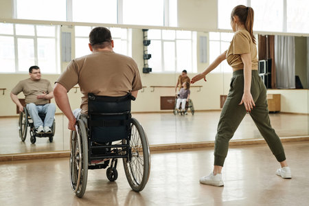 Rear view of man in wheelchair looking at young female dance trainer while she demonstrating moves to himの写真素材