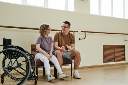 Long shot of cheerful young female wheelchair dancer sitting on bench chatting with her male trainer during breakの写真素材