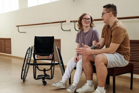 Long shot of modern Caucasian female wheelchair dancer and her partner sitting on bench in dance studio chatting during breakの写真素材