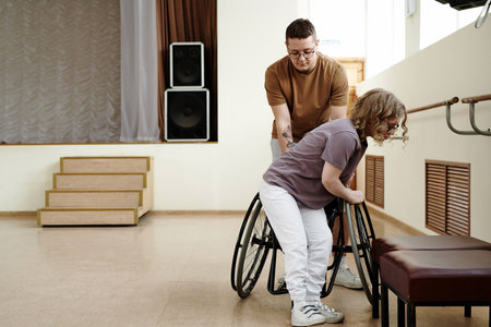 Long shot of young Caucasian man helping woman with disability to get into wheelchair in dance studioの写真素材