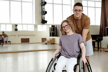 Medium long portarit of cheerful young woman in wheelchair and young man standing behind her in dance studio, copy spaceの写真素材