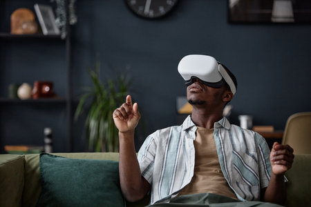Young African American man wearing VR headset sitting on sofa in living room playing video game, copy spaceの写真素材