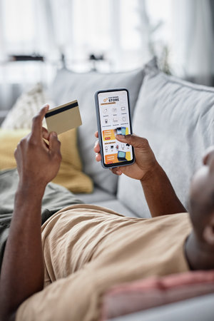 Unrecognizable Black man relaxing on couch in living room at home doing online shopping with use of...