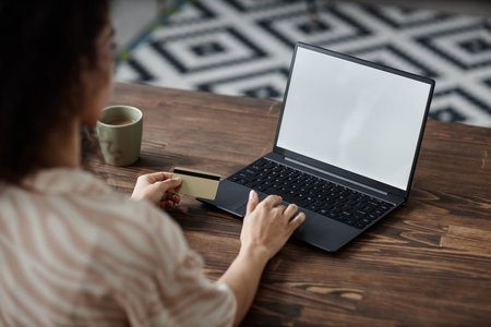 Over the shoulder shot of unrecognizable woman sitting at desk doing online shopping on laptop and paying with bank cardの写真素材