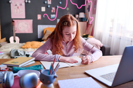 Red-haired teen girl sitting at desk in her room making notes in notebook while doing homework, copy...