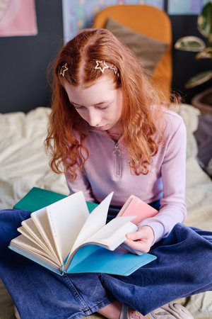 Vertical shot of red-haired Caucasian teen girl sitting on bed reading bookの写真素材