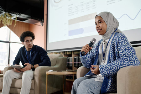 Low angle of young female student wearing hijab sitting in armchair speaking at conference in modern collegeの写真素材
