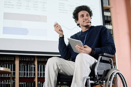 Low angle view of self-confident Middle Eastern student with disability holding digital tablet doing presentation at event in college libraryの写真素材